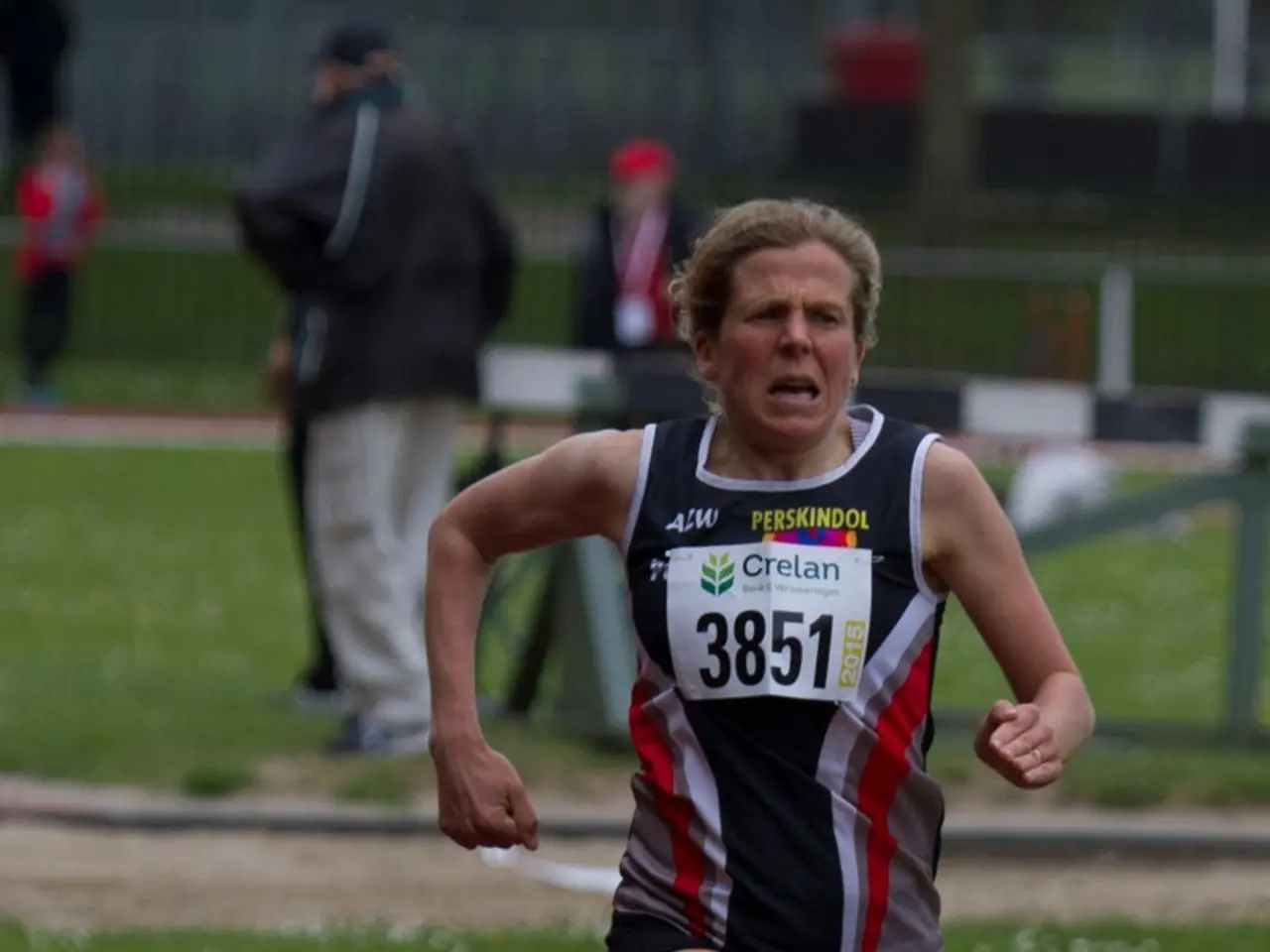 In this image I can see a person running. There is grass and other people at the back.