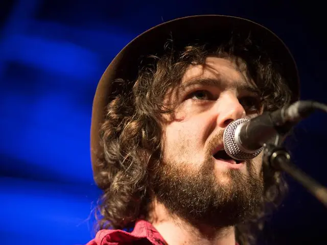 In this picture a man is singing along with playing guitar is wearing a hat. He is wearing a red...
