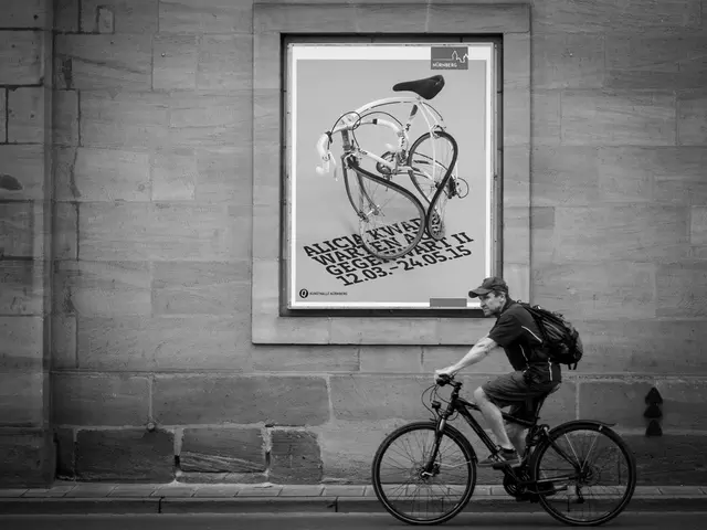There is a man cycling a bicycle on the road. In the background there is an advertisement poster on...