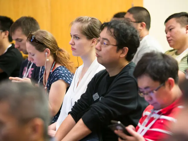 In this image we can see people sitting and some of them are holding mobiles. In the background...