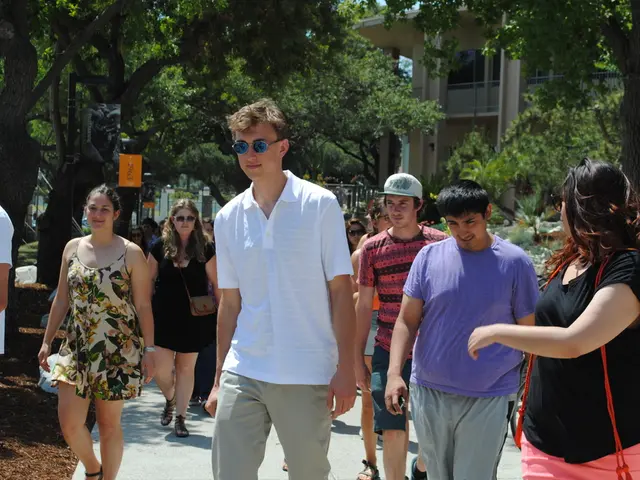 In this image on the path many people are walking. In the background there are trees, buildings.